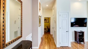 Un pasillo con piso de madera, paredes blancas y un espejo grande a la izquierda. Hay una puerta a la derecha que da a otra habitación, y un televisor montado en la pared sobre ella.