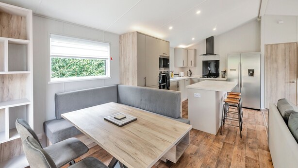 A modern kitchen with built-in appliances, a dining area with a wooden table and grey seating, and a large window with a view of greenery. - Cawood Holiday Home 2 (Cawood, Selby)