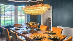 Ein Esszimmer mit großem Holztisch, gedeckt für eine Mahlzeit, umgeben von Stühlen und einer hängenden Kristallleuchter.