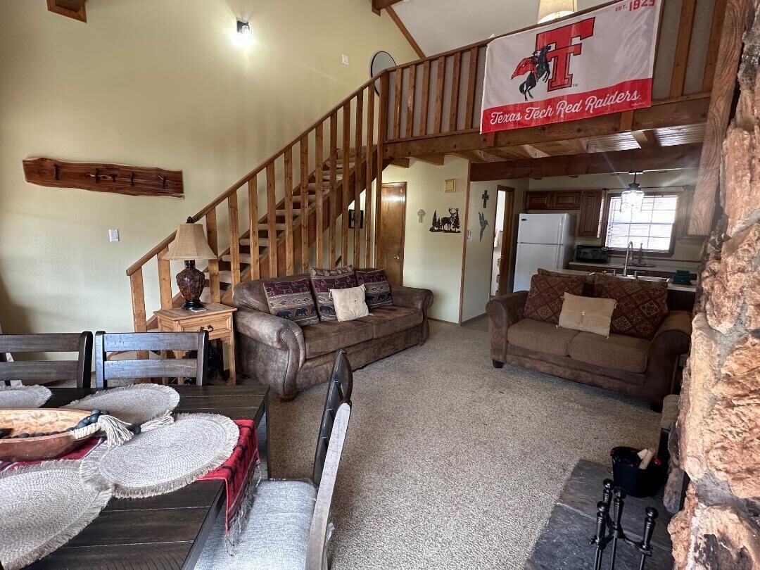 A spacious living area with a wooden staircase, two sofas, a dining table with woven placemats, and a kitchen area visible through an open doorway.