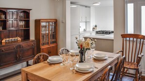 A dining area with a wooden table set for four, surrounded by chairs. A wooden cabinet with glass doors is to the left, and a kitchen area is visible in the background. - 2 bedroom accommodation in Cromer (Cromer)