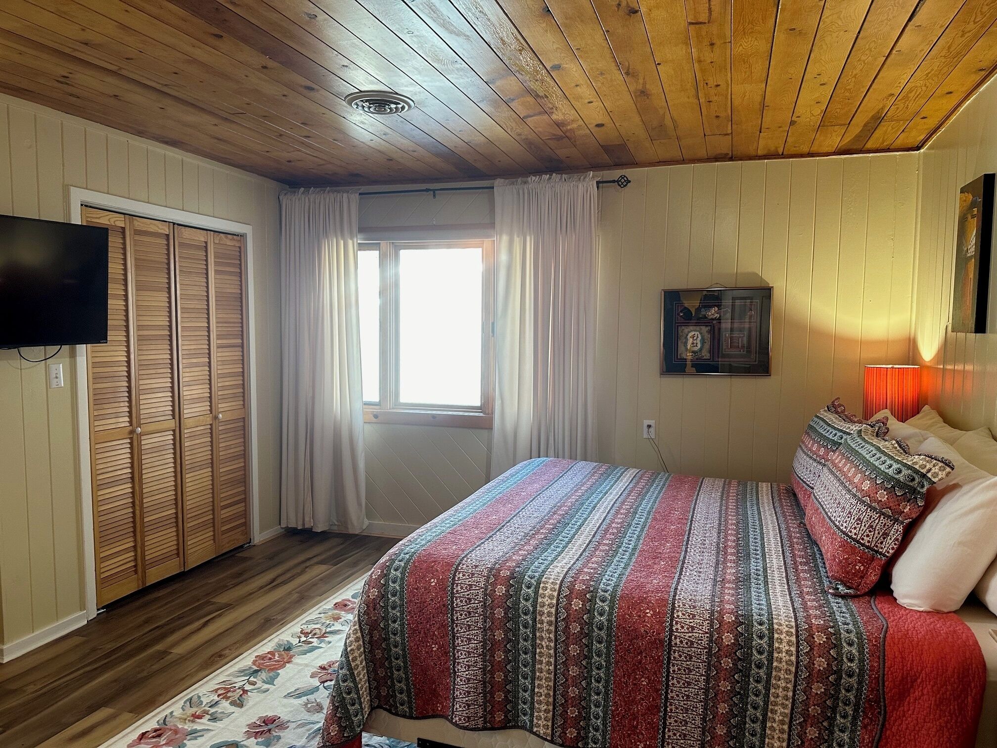 A bedroom with a wooden ceiling, a bed with a patterned bedspread, a television mounted on the wall, and a window with curtains.