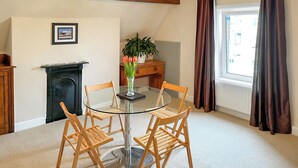 A dining area with a round glass table, four wooden chairs, a vase with flowers, a fireplace, a wooden cabinet, and a window with curtains.