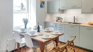 A modern kitchen with white cabinets, a wooden dining table set for four, and a window with a view of buildings. - 3 bedroom accommodation in St Ives (St Ives)