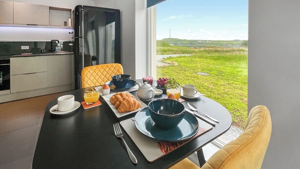 Ein Esstisch mit Croissants, Obst und Kaffee, ein gelber Stuhl und ein Blick auf eine grüne Landschaft durch ein Fenster.
