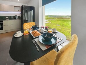 A breakfast table with croissants, fruit, and coffee, a yellow chair, and a view of a green landscape through a window.