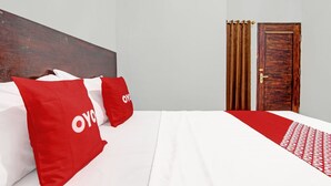 Een hotelkamer met een bed, twee rode kussens, een houten hoofdeinde, een witte spreidek en een kamerkleed met dessin.
