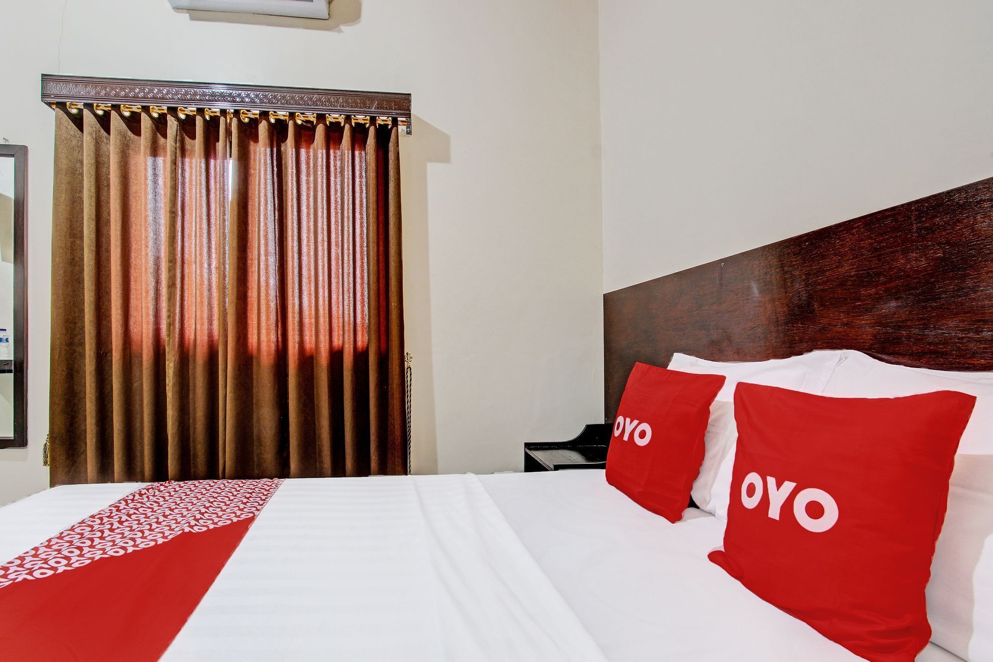 A hotel room with a bed, a headboard, a window with curtains, and a bedside table.