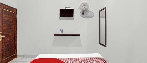 Un dormitorio con cama, una mesita de noche con dos botellas, un ventilador de pared, un televisor pequeño y un espejo.