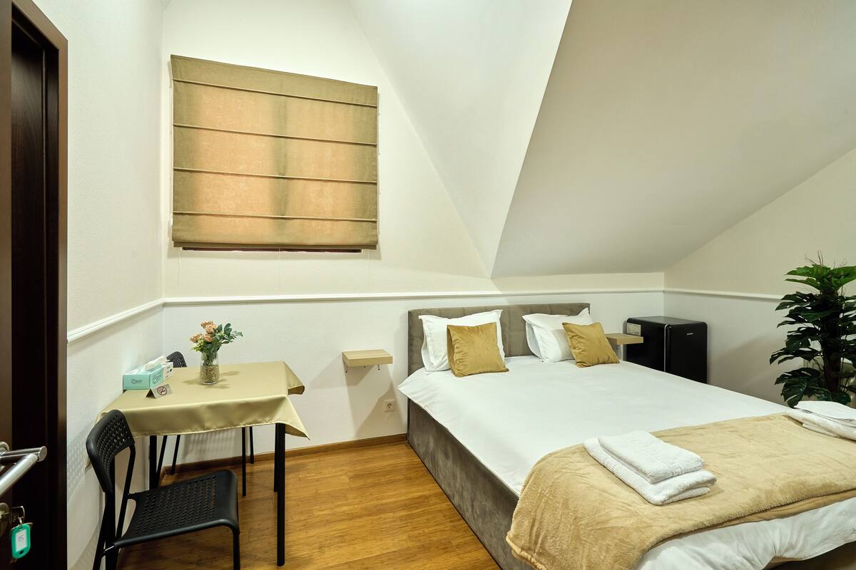 Une chambre à coucher comprenant un lit, une table de chevet, une chaise, une fenêtre et une petite table sur laquelle est posé un vase de fleurs.