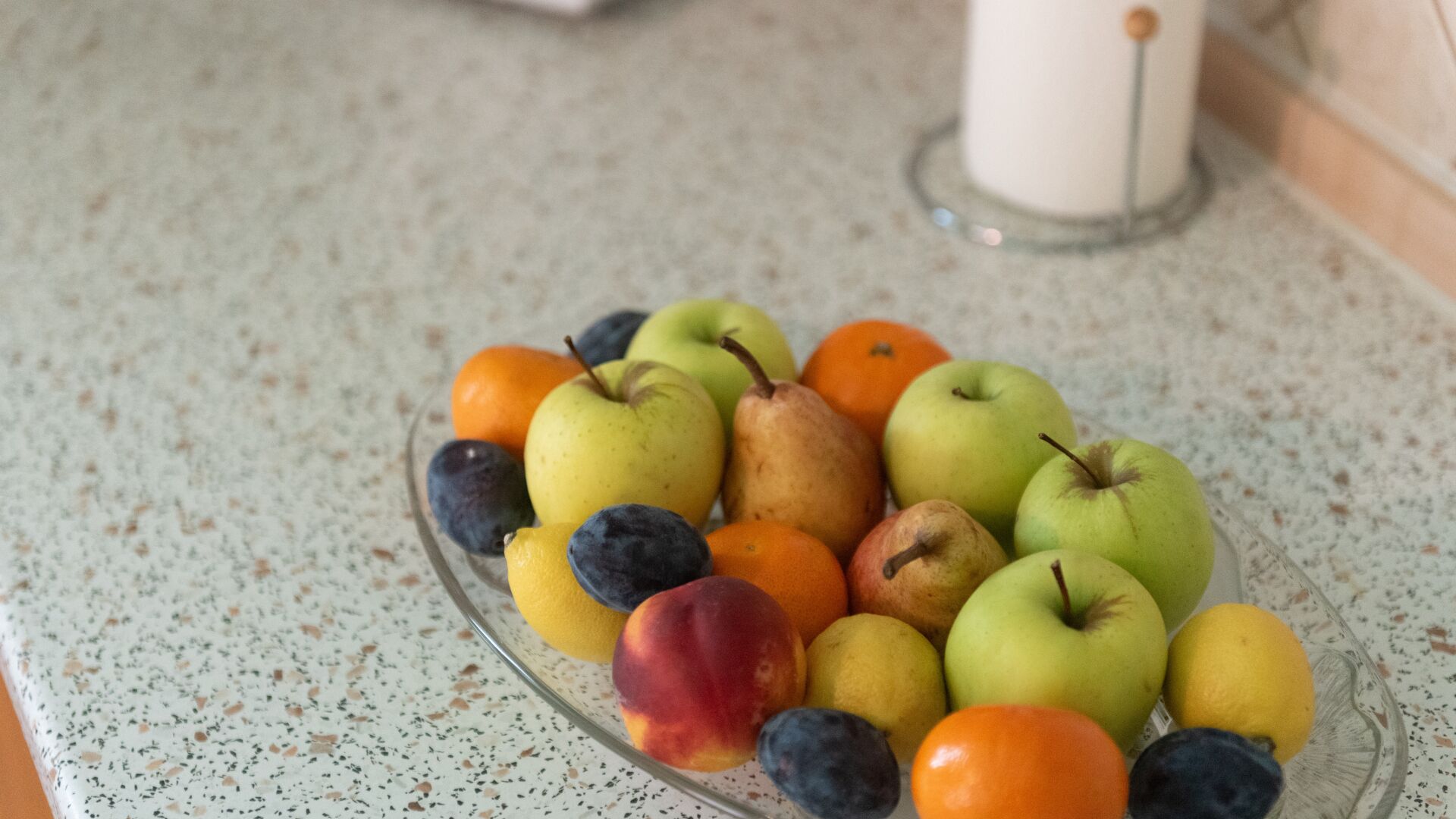 Een glazen schaal met verschillende soorten fruit, zoals appels, sinaasappels en pruimen, op een gekleurd aanrecht.