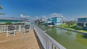 Una terraza de madera con sillas, un canal con casas y un cielo despejado.