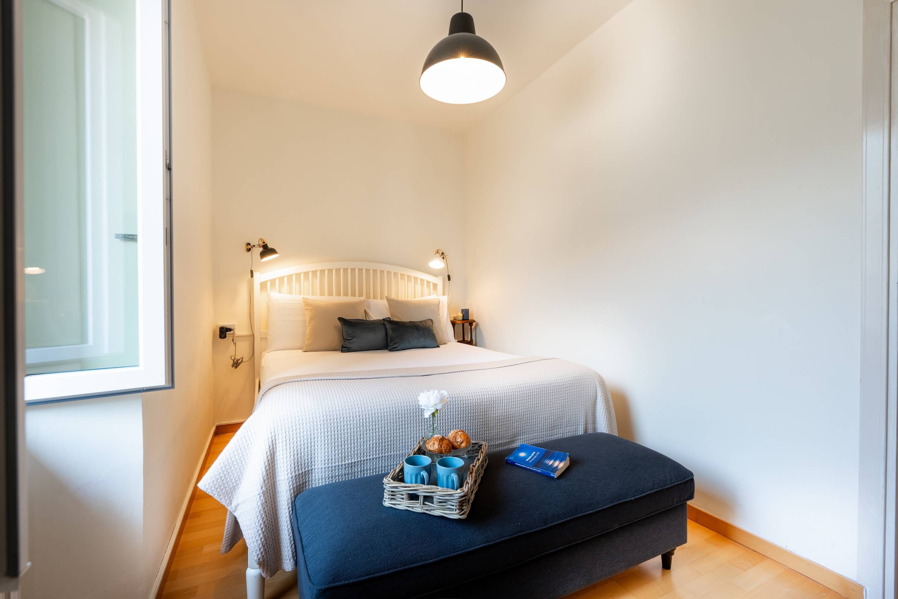 Une chambre à coucher avec un lit, des lampes de chevet, une table de chevet sur laquelle se trouve un panier rempli d’objets, et une fenêtre.