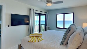 A bedroom with a bed, a ceiling fan, a TV mounted on the wall, and a window with a view of the sea. - Seaside 505 | Top-Floor w. Views +Amenities (Fort Myers Beach)