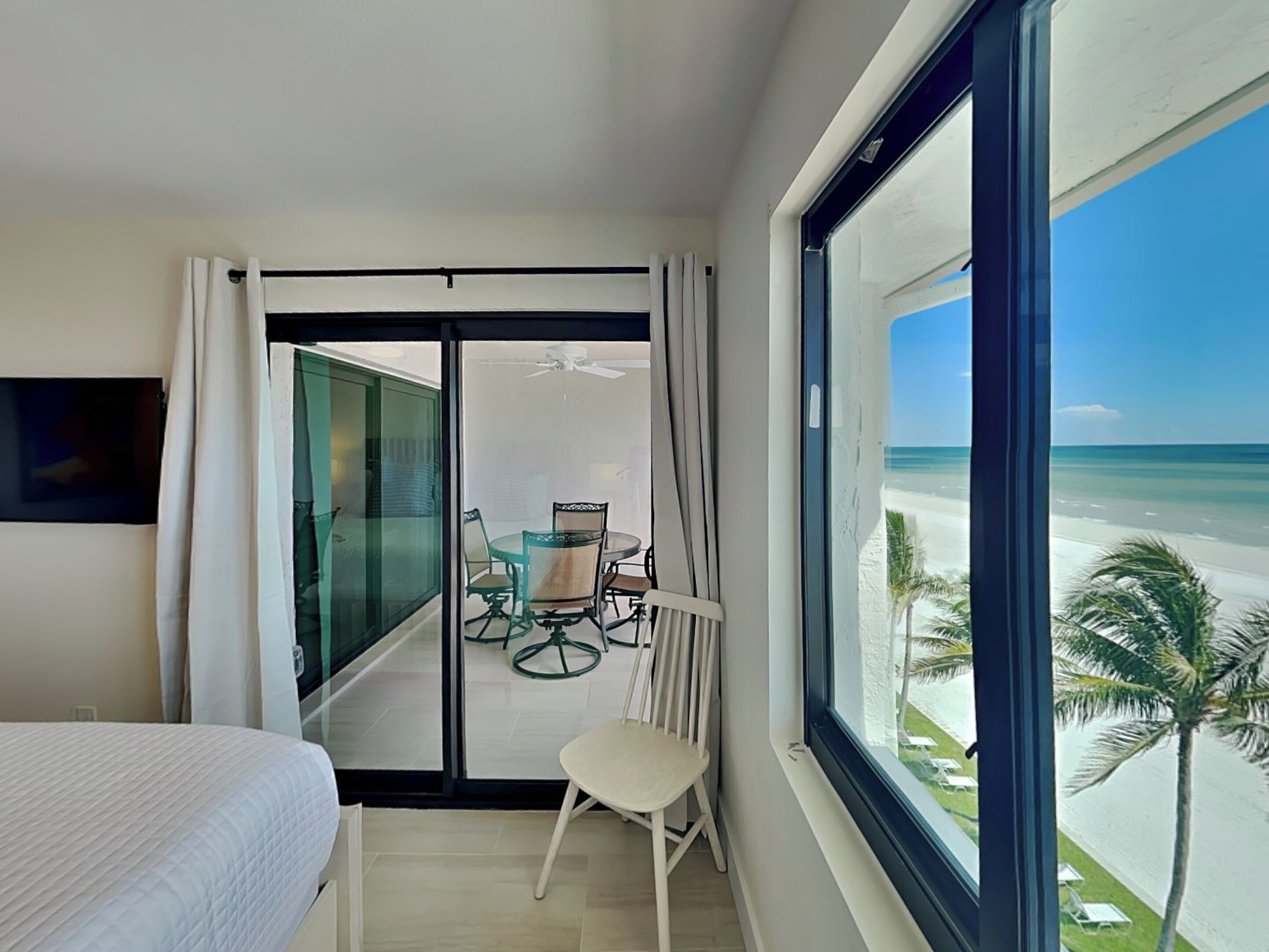 Habitación de hotel con un ventanal grande con vistas a la playa, un balcón con mesa y sillas, y una cama con edredón blanco.