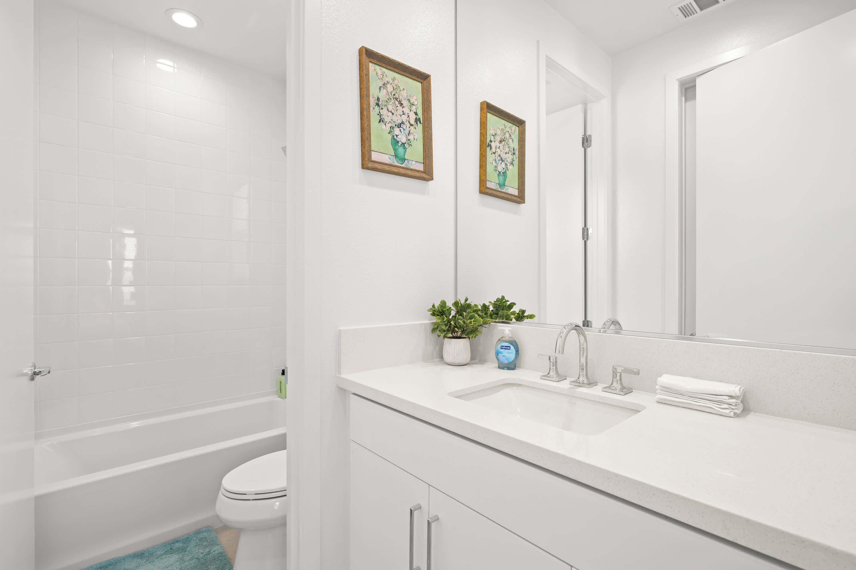 A modern bathroom with a white sink, mirror, and toilet. There are two framed pictures on the wall, a small potted plant, and a blue bottle on the counter.