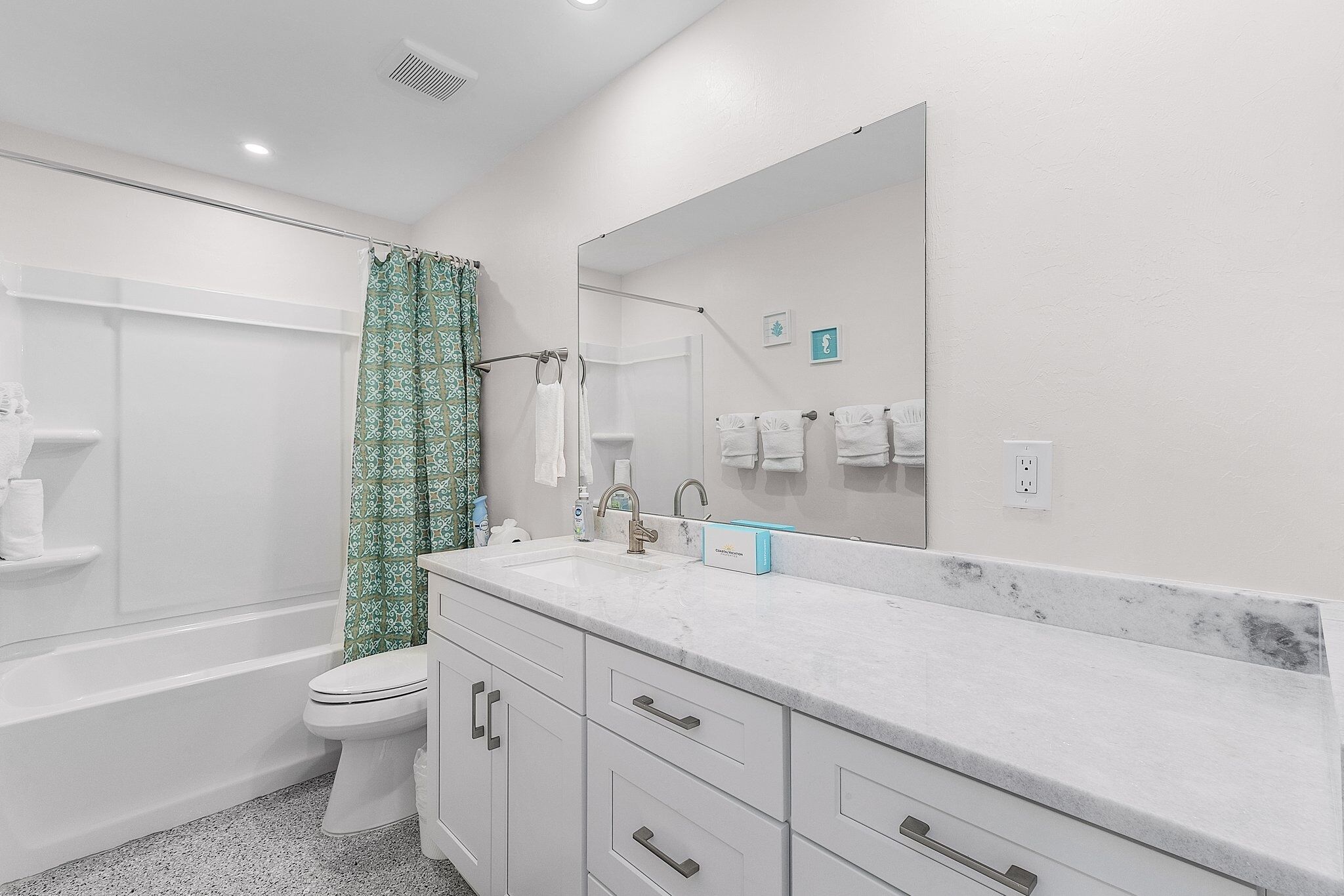 Un baño moderno con un inodoro blanco, una ducha con cortina verde estampada, un espejo grande y una encimera de mármol con dispensador de jabón.