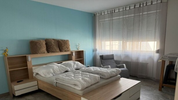 A modern bedroom with a wooden bed, a bedside table, a chair, and a window with blinds. - Horizon 360° (Kitzingen)
