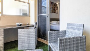Een kamer met een bureau, twee rieten stoelen, een bed en een schuifdeur van glas.
