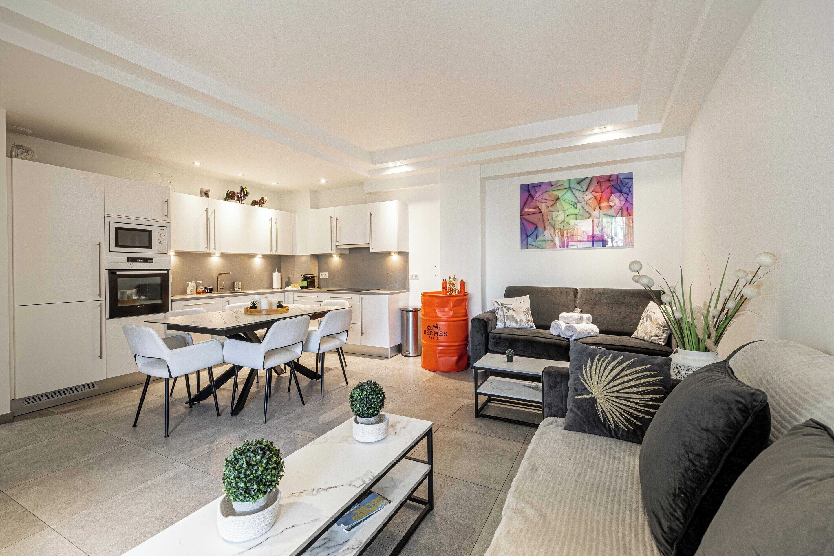 A modern kitchen with white cabinets, a dining area with a table and chairs, a sofa, and a colorful abstract painting on the wall.
