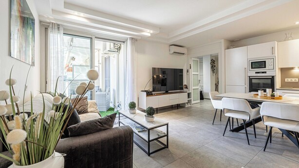 Een moderne woonkamer met een bank, salontafel en een eethoek met stoelen. Er is een televisie en een keuken met kasten en apparatuur.
