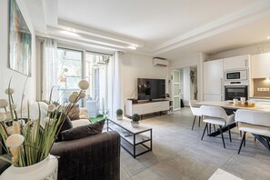 A modern living room with a sofa, coffee table, and a dining area with chairs. There is a TV and a kitchen area with cabinets and appliances. - Modern apt With Balcony - Cannes/croisette (Cannes)