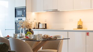 A modern kitchen with a dining table set for two, featuring a vase with dried plants, a loaf of bread, and various fruits. - Gaia Elegant Apartment w/ Patio by LovelyStay (Vila Nova de Gaia)
