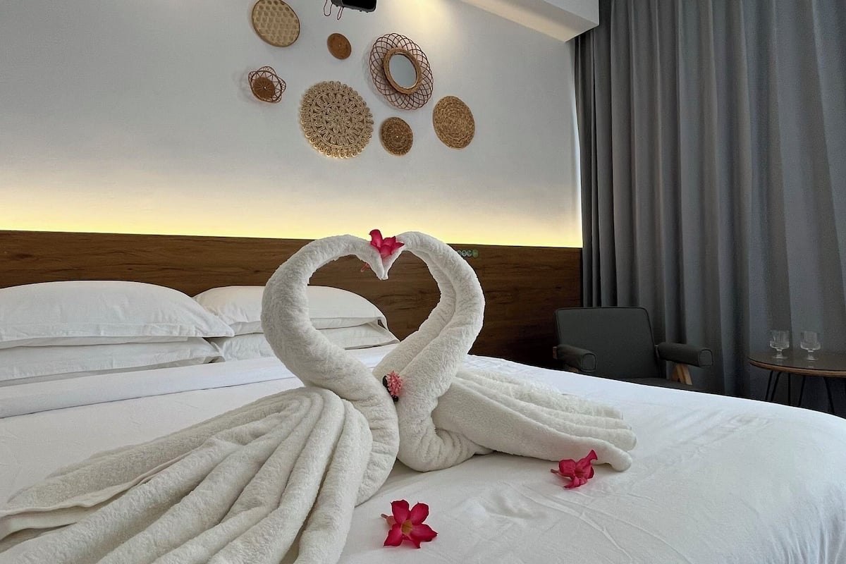 A hotel room with a bed, a wooden headboard, a wall with decorative wall art, a grey curtain, and a small table with glasses.