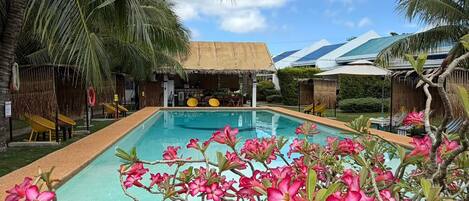 A swimming pool with a tropical garden, palm trees, and a thatched-roof structure.