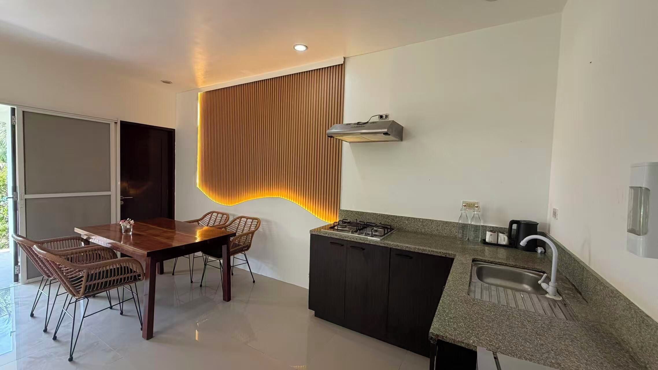 A modern kitchen with a dining area, featuring a wooden table, wicker chairs, a built-in gas stove, and a stainless steel sink.