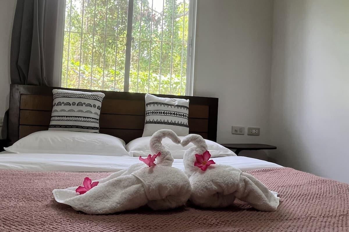A hotel room with a bed, two pillows, a bedside table, and a window with a view of greenery.