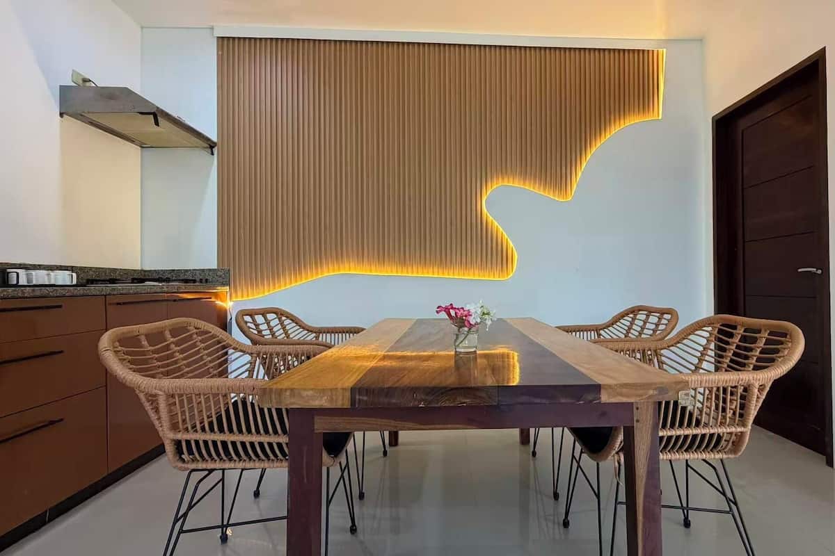 A dining area with a wooden table, wicker chairs, a vase with flowers, and a wall with a vertical light feature.