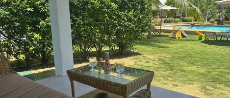 Een terras met een tafel met glazen blad, rieten stoelen en uitzicht op een zwembad en een speeltuin.