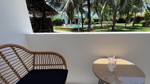 A wicker chair with a black cushion and a small round table with a glass on it, overlooking a pool and palm trees. - Belle Homes (Panglao)