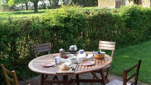 Mesa de madera redonda al aire libre con sillas, rodeada por un seto y árboles.