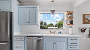 A kitchen with light blue cabinets, stainless steel appliances, and a window with a view of greenery. - BayBreeze Cottage • Family-Friendly • Sleeps 6-8 • Near SRQ, Ringling & Beaches (Sarasota)