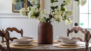 A dining table with a vase of white flowers, surrounded by wooden chairs and plates. - BayBreeze Cottage • Family-Friendly • Sleeps 6-8 • Near SRQ, Ringling & Beaches (Sarasota)