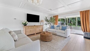 A modern living room with a white sofa, a TV mounted on the wall, a wooden cabinet, and a large window with curtains. - BayBreeze Cottage • Family-Friendly • Sleeps 6-8 • Near SRQ, Ringling & Beaches (Sarasota)