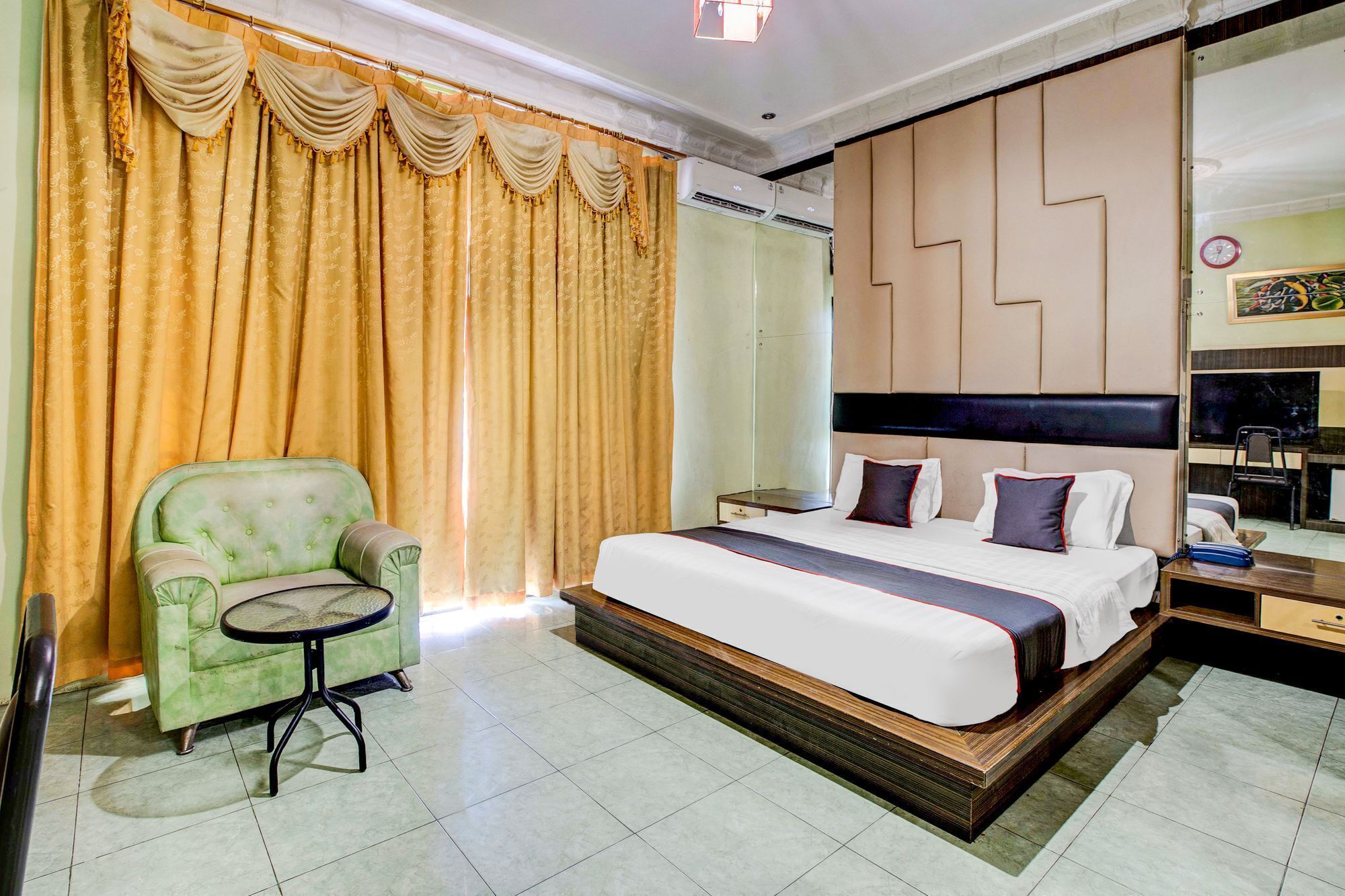 A hotel room with a bed, a green armchair, a small table, and a large mirror.