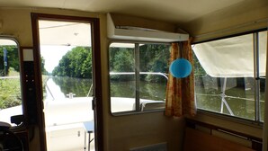 A room with a bed, a window with curtains, a steering wheel, and a view of a river. - Calm overnight aboard the JAVA boat, 30 minutes from PAL, disconnection guaranteed! (Luneau)