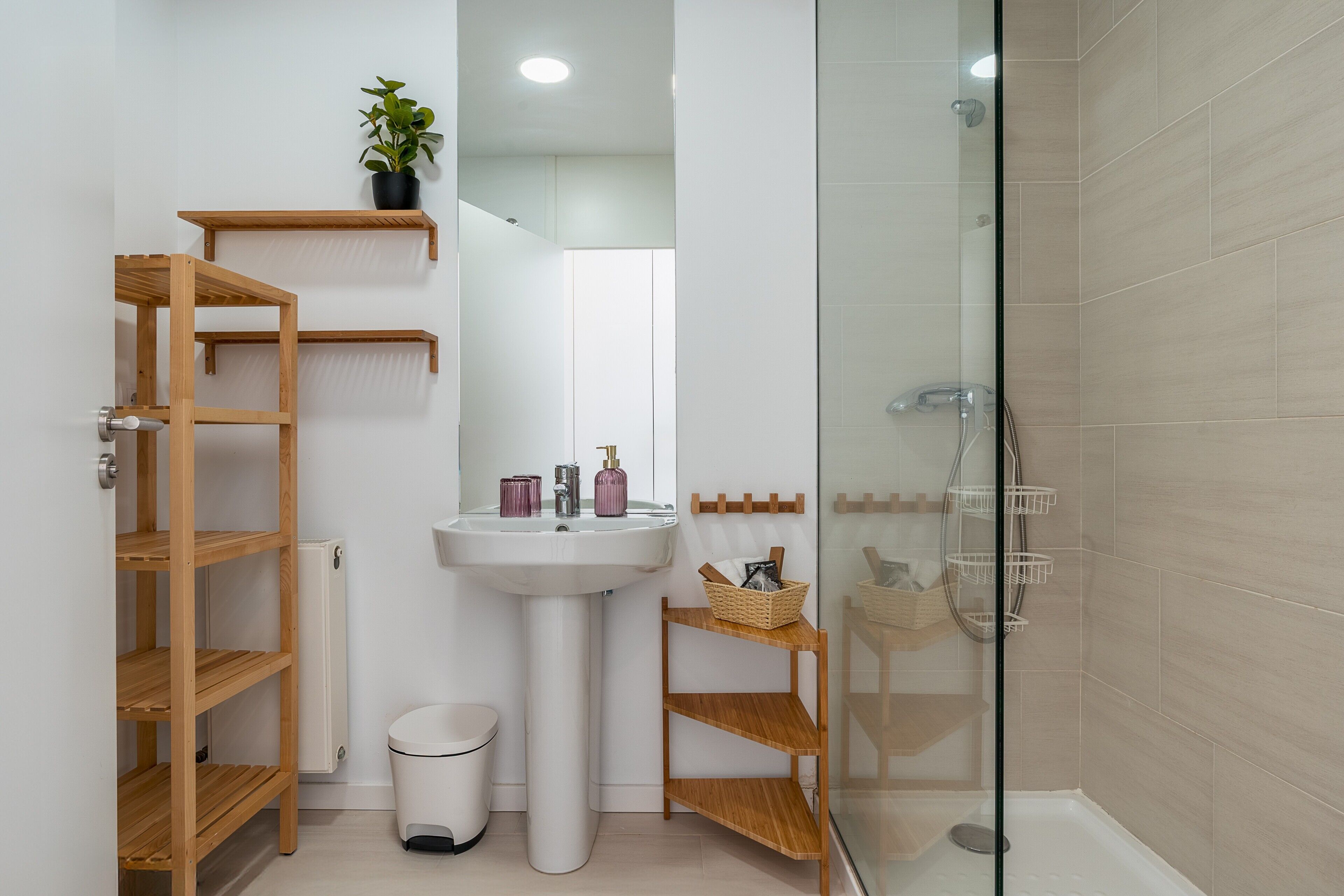A modern bathroom with a glass shower enclosure, a white sink, wooden shelves, and a potted plant.