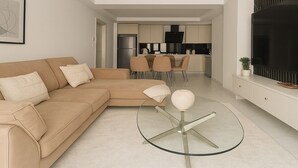 A modern living room with a beige sectional sofa, a glass coffee table, and a TV on a white cabinet. A kitchen with a dining area is visible in the background. - 2 Chambres Spacieux a Dely Ibrahim (Algiers)