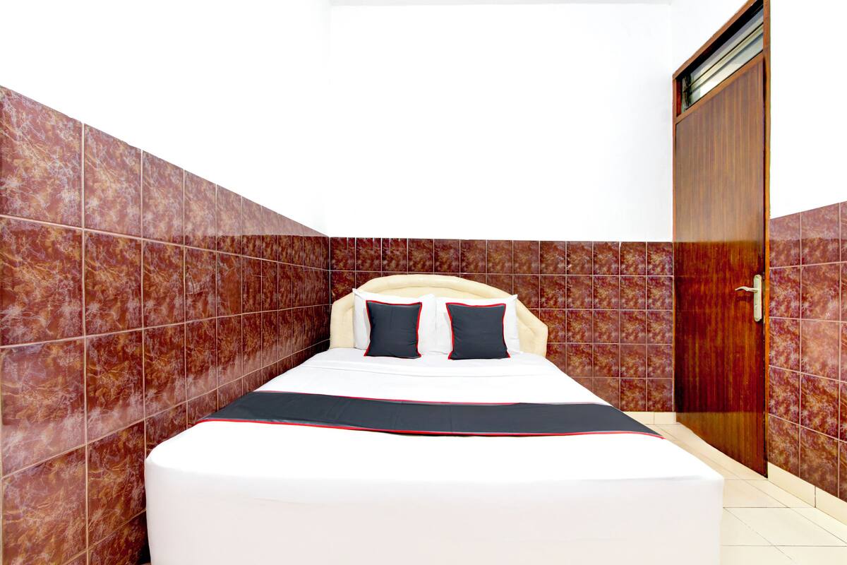 A neatly made bed with a white and red bedspread, two black pillows, and a wooden wardrobe in a room with red-tiled walls.