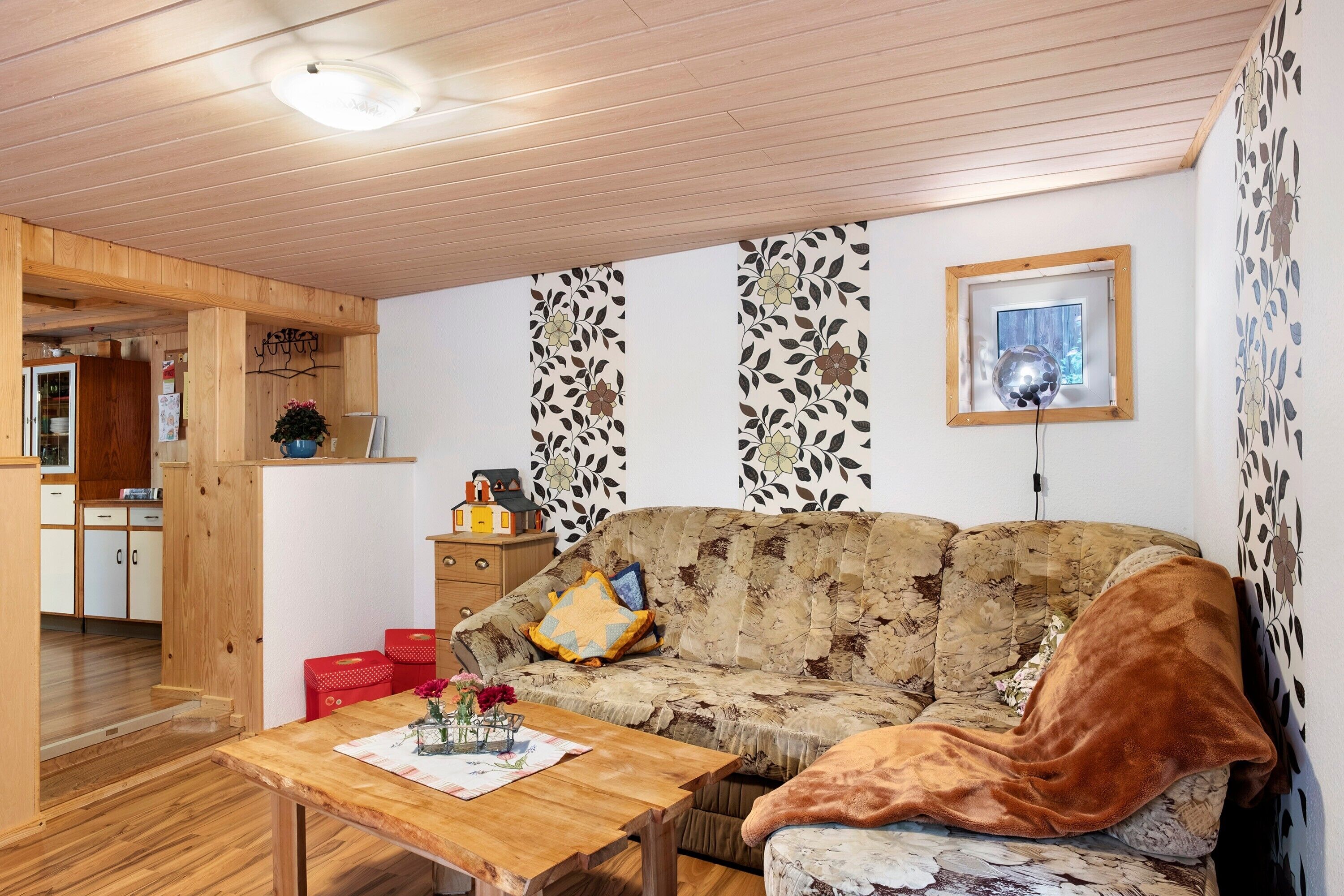 Una sala de estar acogedora con techo de madera, un sofá con cojines estampados, una mesita con un florero y una ventana con un marco decorativo.