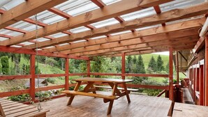 A covered outdoor seating area with wooden benches and picnic tables, large windows, and a view of a green landscape. - Apartment 'Schwäblehof' (Triberg im Schwarzwald)