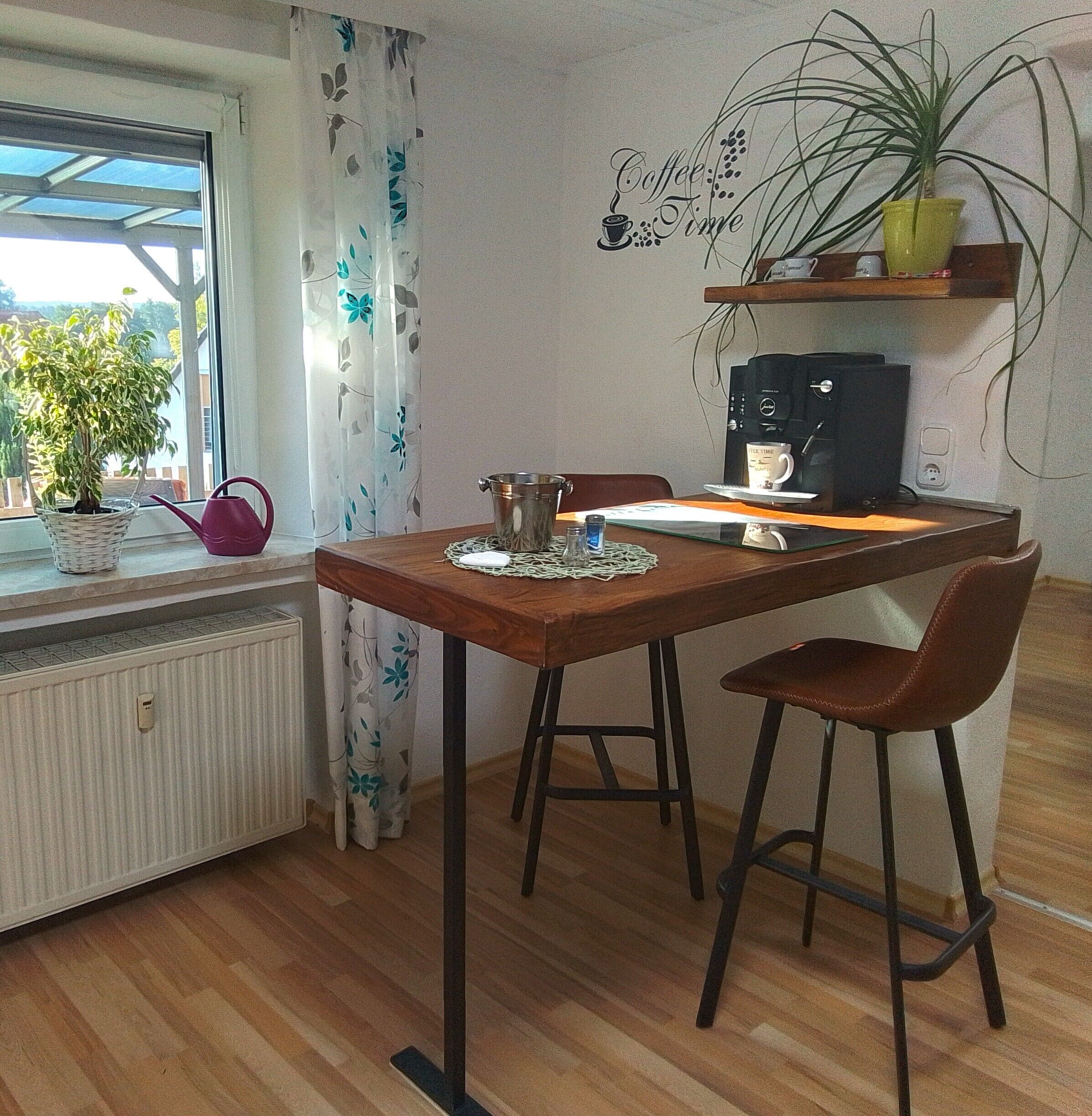 Un comedor con una mesa de madera, dos sillas, una cafetera, una planta en maceta y una ventana con cortinas.