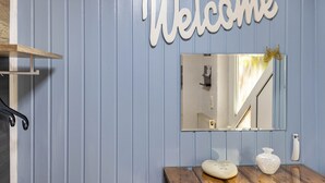 Un baño con un letrero que dice 'Welcome', un espejo, un tocador de madera y un pequeño florero.