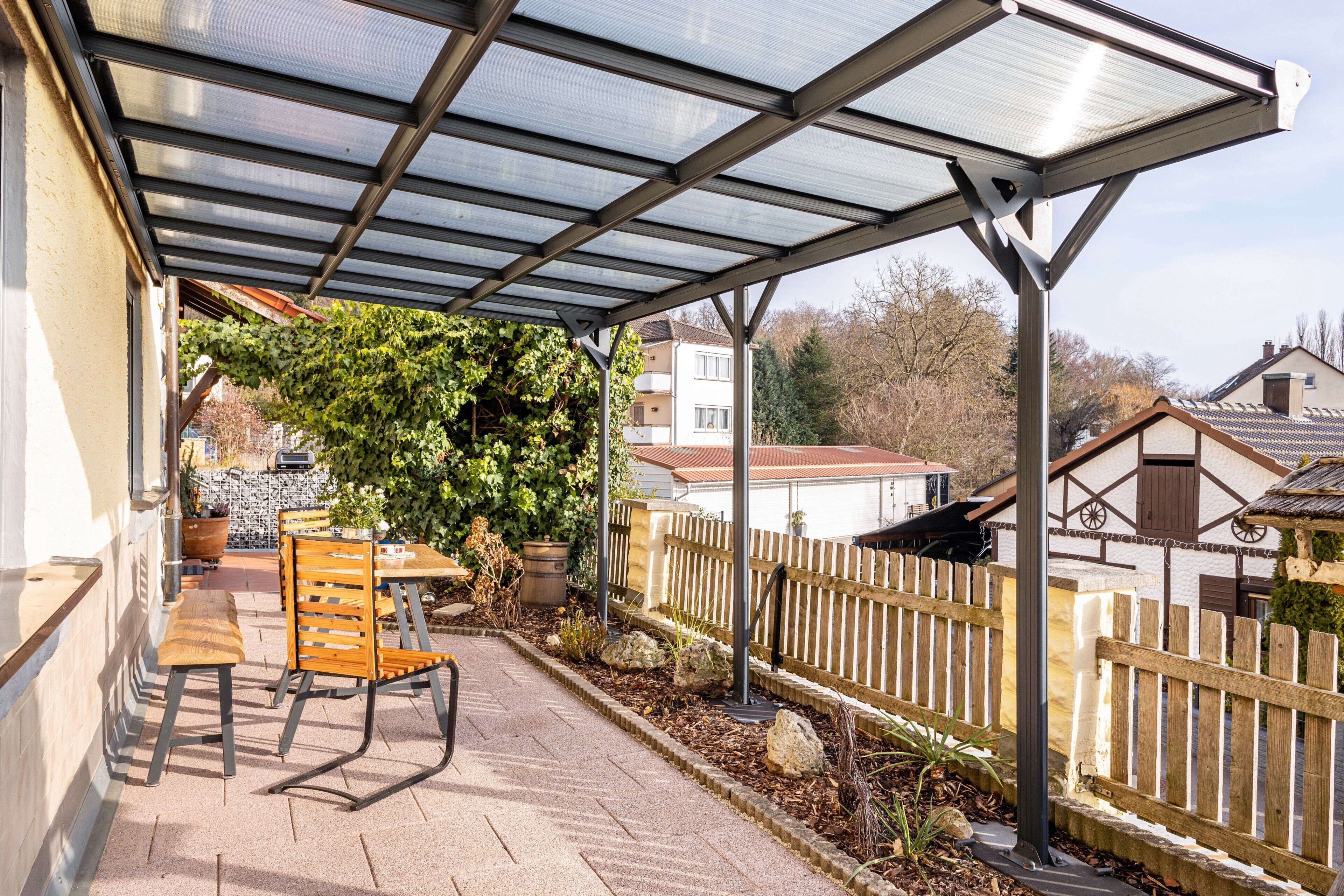 A covered patio with a table and chairs, a wooden fence, and a house in the background.