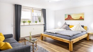 Un dormitorio con una cama de madera, un sofá gris, una mesita y una ventana con cortinas.