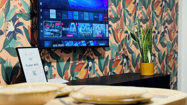 A hotel reception area with a flat-screen TV displaying various streaming services, a 'Guest Wifi' sign, and a plant on a wooden table. - Vimini Like a Local (Bari)